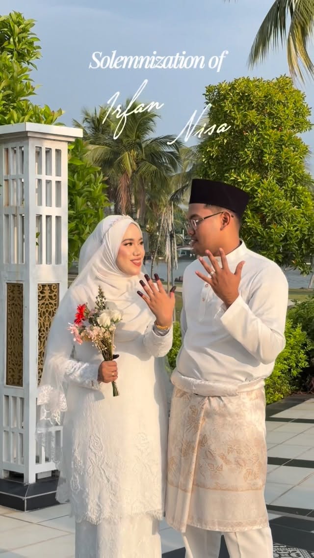 A couple in cream outfits holding up their hands outdoors beside a white gate