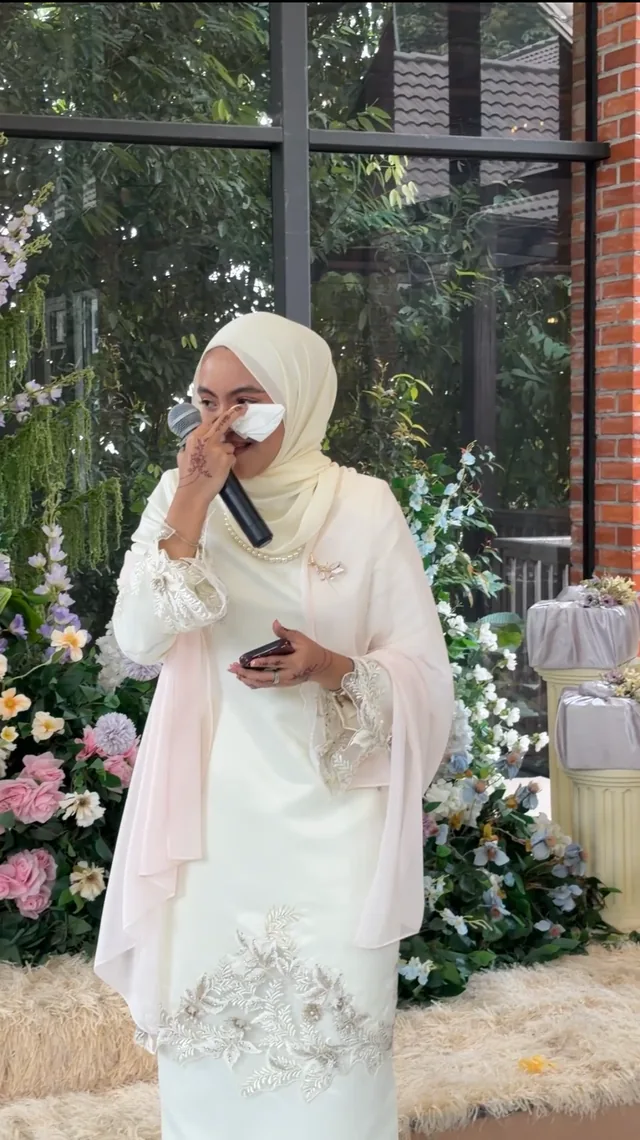 A woman in white holding a tissue to her eye beside flowers and a window